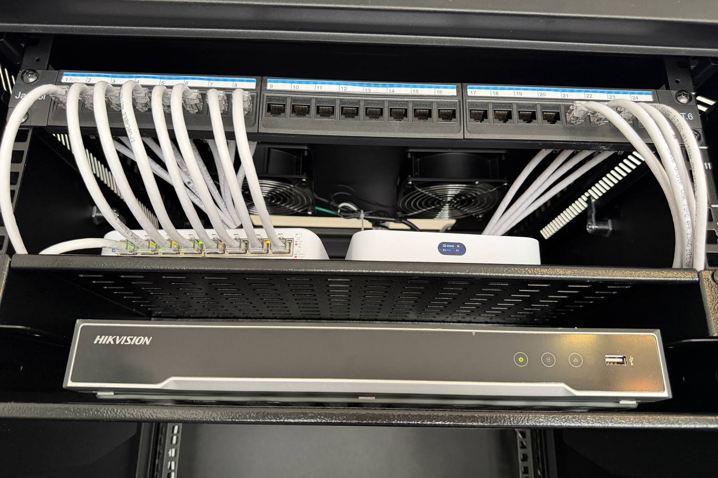 A network rack with a labeled patch panel, white patch cables, a network switch and gateway, and a camera recorder on the shelf below